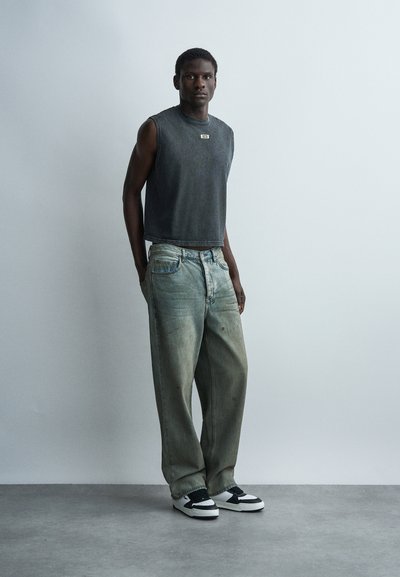 Άνδρας που στέκεται φορώντας αμάνικο σκούρο γκρι τοπ, φαρδιά ξεθωριασμένα μπλε τζιν και ασπρόμαυρα αθλητικά παπούτσια σε απλό ανοιχτό φόντο.