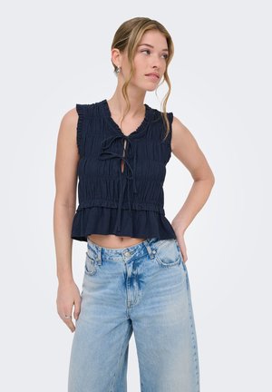 Jeune femme aux cheveux clairs portant un haut noir sans manches froncé avec des liens et un jean bleu clair taille haute, regardant sur le côté.