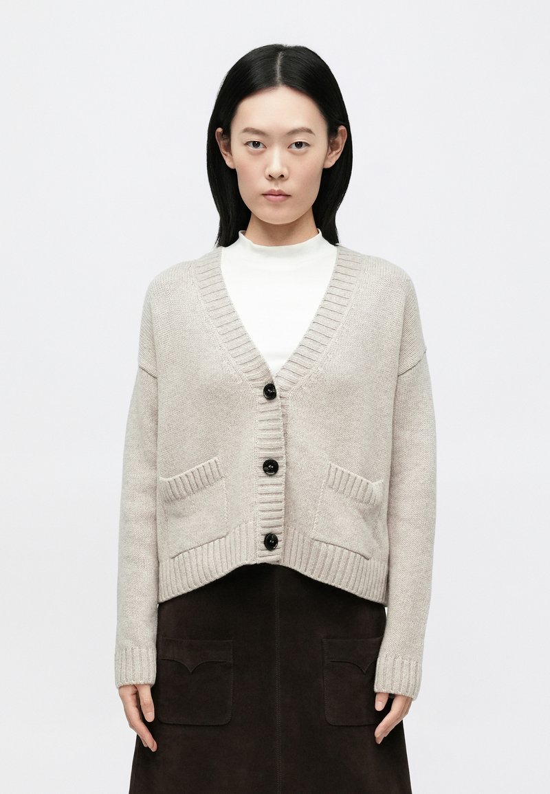 Cardigã bege em malha com decote em V, punhos canelados e dois bolsos frontais. Apresenta botões pretos e uma bainha curta.