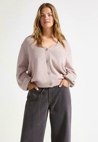 Cardigã de malha cor de rosa claro com decote em V, fecho de botões e bainha canelada. Combinado com calças largas cinza-escuro folgadas.