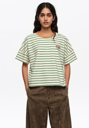 Mujer con cabello largo y oscuro, vestida con una camiseta blanca de manga corta con rayas verdes y pantalones marrones de pierna ancha, de pie con las manos en los bolsillos.