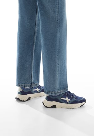 Navy blue sneakers with white logo, designed with a mix of textured materials and a lightweight sole, paired with wide-leg light blue jeans.