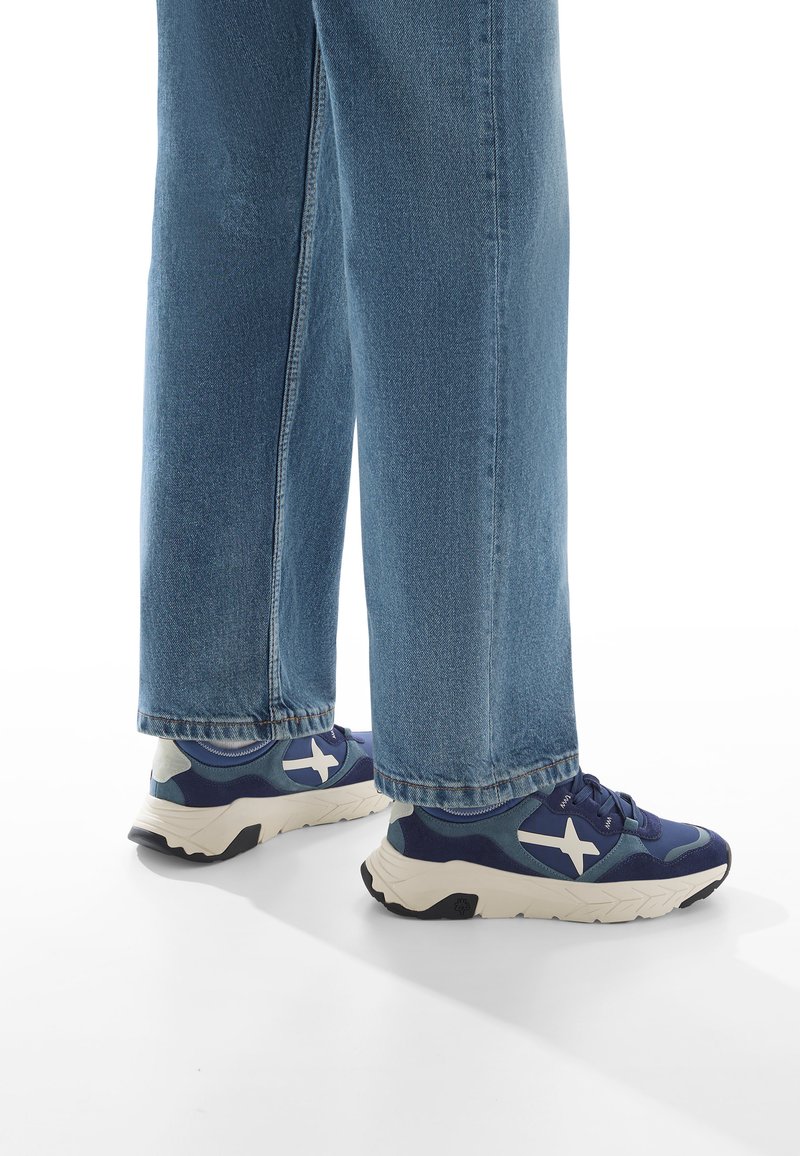 Baskets bleu marine avec logo blanc, conçues avec un mélange de matériaux texturés et une semelle légère, associées à un jean bleu clair à jambes larges.