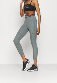 Harmaat korkeavyötäröiset legginsit, joissa on teksturoitu pinta, varustettu Nike-logolla vyötäröllä ja tyylikkäällä muotoilulla, yhdistetty mustiin urheilukenkiin.