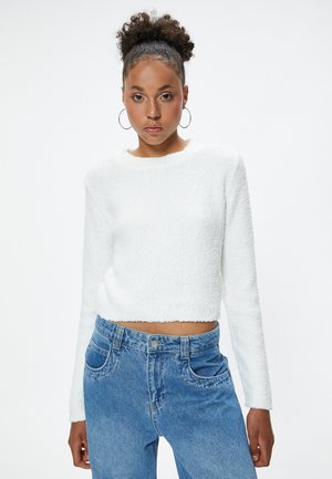 Pull blanc coupé avec un tissu texturé, des manches longues et un ourlet brut, associé à un jean bleu taille haute avec une fermeture à boutons visible.