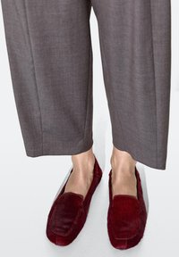 Mocassini in pelliccia bordeaux con una texture morbida e bordi arrotondati, abbinati a pantaloni larghi grigi a vita alta.