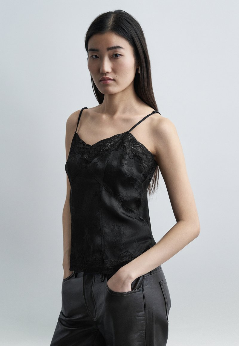 Νεαρή γυναίκα με μακριά σκουρόχρωμα μαλλιά φοράει μαύρη κορυφή με δαντέλα και μαύρο δερμάτινο παντελόνι, τα χέρια στις τσέπες, ουδέτερο γκρι φόντο.