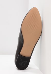 Chaussure plate noire à bout pointu inclinée vers le haut, montrant une semelle lisse couleur camel et un talon texturé avec un logo papillon.