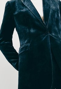 Blazer en velours vert foncé avec fermeture à un bouton et poche à rabat, porté sur un haut blanc.