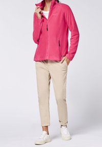 Pink fleece zip-up jacket with high collar, two side pockets, and black zipper details. Beige pants and white sneakers complete the outfit.