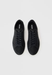 Black sneakers with a textured finish, featuring a rounded toe, flat sole, and tonal laces. Branding visible on the inner sole.