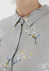 Lichtgrijze blouse met een kraag, voorzien van een bloemenprint in wit, geel en blauw. Gladde textuur met knoopsluiting.