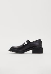 Black leather shoe with a rounded toe, cutout design, and velcro strap. Features a chunky heel and textured rubber sole for traction.