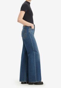 Personne portant un jean bleu taille haute à jambes larges, un haut noir à manches courtes et des chaussures noires, debout avec une main dans la poche sur fond blanc.