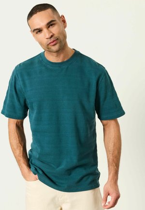 Man with short hair wearing a textured teal short-sleeve shirt and light beige pants, standing with one hand in pocket.