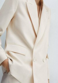Blazer beige de doble botonadura con bolsillo de solapa, llevado por una persona con la mano metida en el bolsillo de pantalones beige a juego.