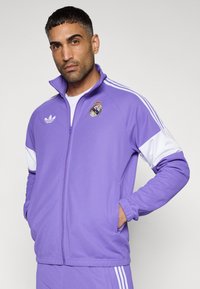 Μωβ σακάκι με φερμουάρ, κατασκευασμένο από συνθετικό ύφασμα, με λευκές λεπτομέρειες, ένα λογότυπο και τρεις λευκές ρίγες στα μανίκια. Ριμπ μανσέτες και τελείωμα.