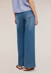 Jeans de denim azul de pierna ancha con un lavado desgastado y detalles de costura visibles, combinados con sandalias marrones. La vista muestra el perfil trasero y lateral.