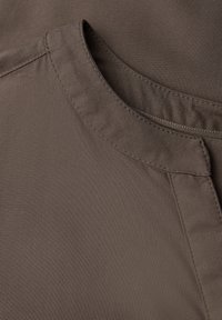 Brunt stof med en blød tekstur, der har en rund halsudskæring og syede detaljer langs kanterne. Nærbillede fremhæver materialets kvalitet.