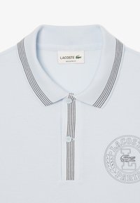 Ljust blå polo skjorta i texturerad tyg, med en svart randig krage, två knappar och en broderad logga på bröstet.