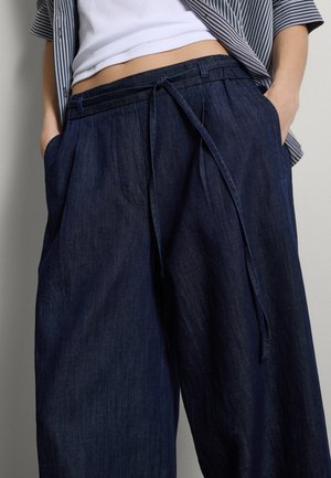 Person trägt weite dunkelblaue Jeanshosen mit Kordelzug in der Taille und Händen in den Taschen, kombiniert mit weißem Top und gestreiftem Hemd.