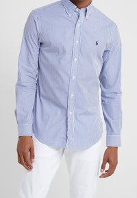 Blå och vit randig skjorta med knappar, krage, långa ärmar och en liten logotyp på bröstet. Tyget ser slätt och lätt ut.