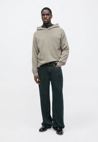 Světle šedá žebrovaná mikina s kapucí a přední kapsou, v kombinaci s volnými tmavými džínami a černou obuví. Jednoduchý design s volným střihem.