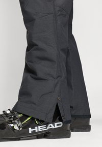 Mustad suusapüksid tekstureeritud viimistlusega, külgmiste lukkudega ja nöppidega pahkluude juures, kantud mustade suusasaabaste kohal, millel on rohelised aktsendid.