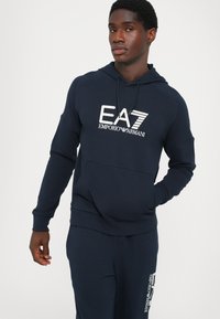 Felpa con cappuccio blu navy realizzata in tessuto morbido, dotata di una tasca frontale e logo EA7 Emporio Armani stampato in bianco.