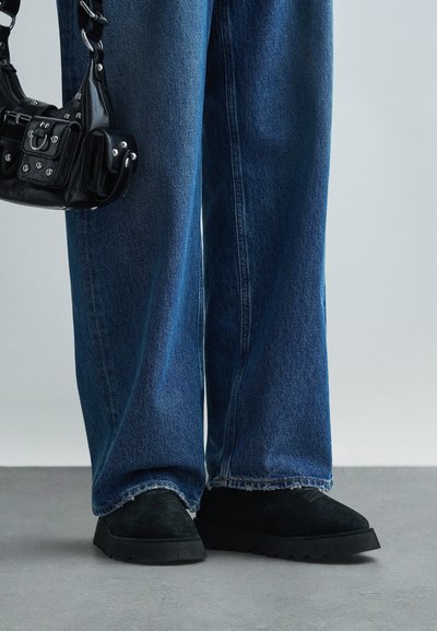 Czarne zamszowe buty z solidną podeszwą w połączeniu z niebieskimi spodniami o szerokich nogawkach z postrzępionymi końcami, na neutralnym tle.