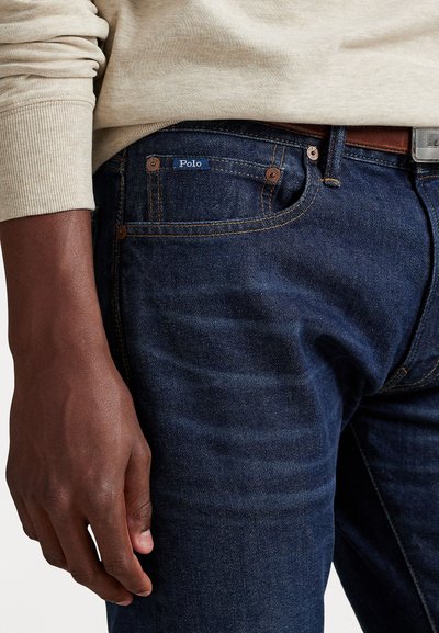 Dark blue denim jeans with orange stitching, a small blue "Polo" logo, and copper-tone hardware. The hand rests on the hip.
