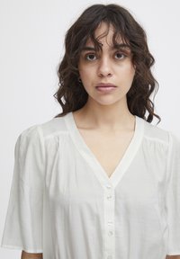 Blouse blanche légère avec des manches bouffantes, décolleté en V et une rangée de boutons sur le devant ; texture lisse et léger éclat.
