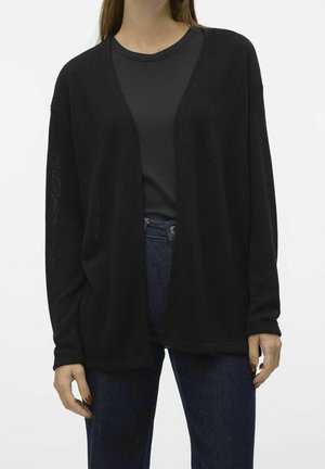 Person trägt schwarzen Cardigan mit offenem Vorderteil über dunklem, geripptem Oberteil und blauer Jeans, steht vor schlichtem hellem Hintergrund.