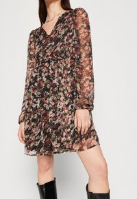 Kukkamekko läpinäkyvästä mustasta kankaasta, jossa on pitkät hihat, v-kaula-aukko ja istuva vyötärö. Värit sisältävät vaaleanpunaisia, punaisia ja vihreitä kukkakuoseja.