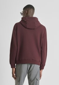Felpa bordeaux con un ampio cappuccio e polsini a costine, realizzata in un tessuto morbido, indossata con pantaloni cargo grigi, mostrando un design semplice ma moderno.