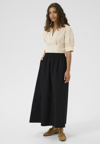 Göndör hajú nő krémszínű, puffos ujjú blúzt, fekete, bő szárú nadrágot és bézs loafert visel, egy kezével a zsebében áll.