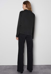 Schwarze Bluse mit langen Ärmeln und Kragen, mit glatter Textur, lockerer Passform und seitlichen Schlitzen. Kombiniert mit hochtaillierten schwarzen Bootcut-Hosen.