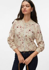 Femme portant une veste bomber zippée beige à motifs floraux avec des poignets côtelés, associée à un jean bleu foncé, tenant une anse de sac marron.