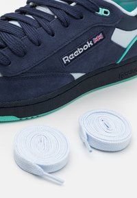 Námořnické semišové tenisky s bílými a tyrkysovými akcenty, s texturovaným povrchem, modrými tkaničkami a logem Reebok. Součástí jsou také dva světle modré tkaničky.