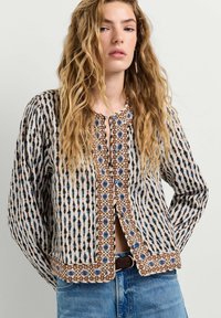 Vrouw met lang golvend blond haar draagt een blouse met patroon en knopen en een blauwe spijkerbroek, staand tegen een effen lichte achtergrond.