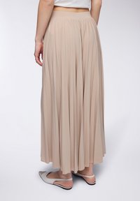Gonna a pieghe lunghezza caviglia in tessuto beige, con una vita larga. La texture è liscia, con un design fluido che si adagia elegantemente.