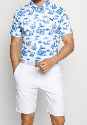 Camiseta polo blanca con un patrón de escena pastoral azul que presenta animales y humanos. Se lleva con pantalones cortos blancos y una hebilla de cinturón plateada.
