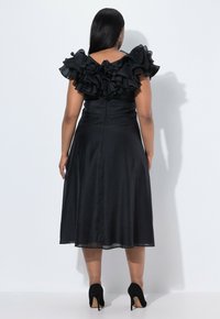 Robe noire avec des manches à volants en décolleté, corsage ajusté et jupe évasée. Tissu lisse avec un léger éclat, portée avec des talons hauts noirs.