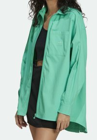 Mujer con una chaqueta holgada color verde menta con cremallera y bolsillo en el pecho, y una parte superior negra recortada junto con una falda debajo.