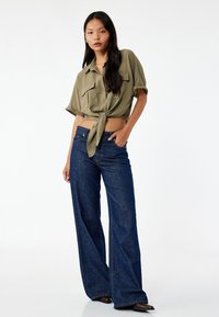 Olivengrøn cropped button-up skjorte med knyttet front og opprullede ærmer, parret med mørkeblå vidde jeans og spidse sorte sko.