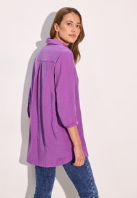 Camicetta viola a maniche lunghe con colletto, vestibilità rilassata e dettaglio a pieghe sul retro. Abbinata a jeans di denim. Tessuto dalla texture liscia.