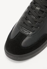Mustad sneaker, mis on valmistatud sileda nahast ja pehmest sametist, ringjate paeltega, madala profiiliga ning peente õmblustähtede detailidega.