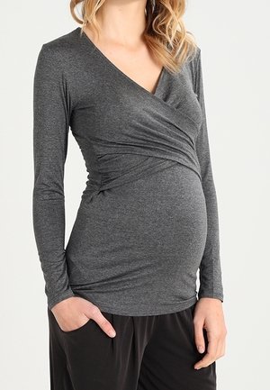 Femme enceinte portant un haut cache-cœur à manches longues gris foncé et un pantalon noir, une main dans la poche sur un fond uni.