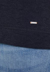 Mörk marinblå textil med liten metalletikett som säger "BOSS HUGO BOSS" ovanför ljusblå denimtyg.