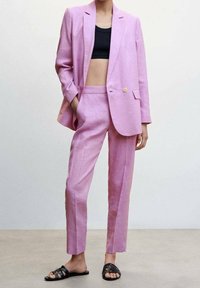 Jasnoróżowy garnitur z lnu, składający się z luźnej marynarki i prostych spodni, zestawiony z czarnym topem na krótki rękaw oraz czarnymi sandałami na cienkich paskach.
